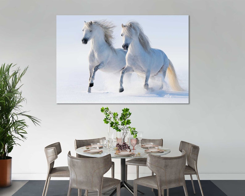 May include: Two white horses running through a snowy field. The horses are running towards the right side of the image. The snow is falling softly and the sky is a light blue.