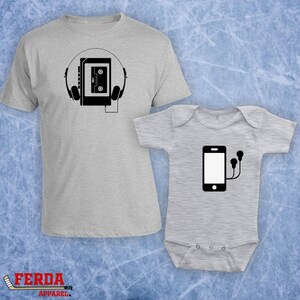 Pode incluir: Um conjunto de duas camisetas cinzas, uma para adulto e outra para bebê. A camiseta para adulto apresenta uma ilustração em preto e branco de uma fita cassete com fones de ouvido. A camiseta para bebê apresenta uma ilustração em preto e branco de um smartphone com fones de ouvido.