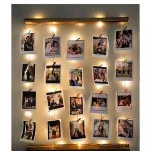 Può includere: Un espositore fotografico in legno con luci a fatina. Il display presenta più foto di famiglia attaccate a corde, illuminate da luci bianche calde. La cornice in legno aggiunge un tocco rustico all'arredamento della parete.