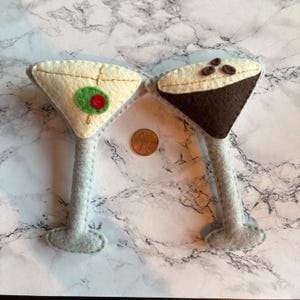May include: Two felt martini ornaments. One is cream-colored with a green olive and red center, the other is brown with coffee beans. Both have gray stems and bases. A penny is between them.