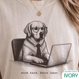 Peut inclure: T-shirt ivoire avec une illustration sépia d'un chien portant des lunettes et une cravate, travaillant sur un ordinateur portable. Le texte "work hard. fetch later." est imprimé en dessous. Le t-shirt est de Comfort Colors.