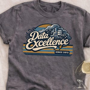 Puede incluir: Camiseta gris con un diseño gráfico que dice "Data Excellence" en una fuente estilizada. El diseño incluye imágenes relacionadas con datos, como gráficos y nubes, con el texto "Since 2012" debajo. La camiseta tiene un aspecto vintage y desgastado.