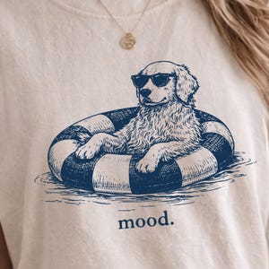 May include: An off-white t-shirt featuring a dark blue graphic illustration of a dog wearing sunglasses while floating in a striped inner tube. Below the graphic, the word mood. is printed in dark blue lowercase letters.