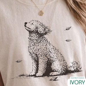 Könnte beinhalten: Ein elfenbeinfarbenes T-Shirt mit einer dunkelbraunen Strichzeichnung eines lockigen Hundes, der im Profil sitzt und den Kopf nach oben neigt. Mehrere kleine, einfache Blattumrisse sind um den Hund mit dem strukturierten Fell verteilt.