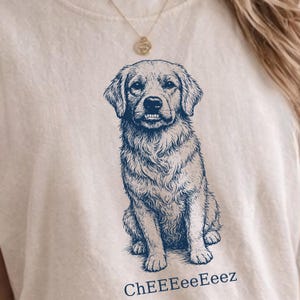 Puede incluir: Camiseta color crema con una ilustración azul detallada de un perro Golden Retriever. El perro está sentado y sonriendo, con el texto "ChEEeEeEeez" impreso debajo. Un collar dorado es visible en la parte superior.