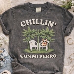 Könnte beinhalten: Dunkelgraues T-Shirt mit dem weißen Schriftzug "CHILLIN' CON MI PERRO". Die Grafik zeigt einen Hund mit Sonnenbrille, der sich in einem Stuhl unter Palmen entspannt. Das Shirt hat einen Vintage-Look.