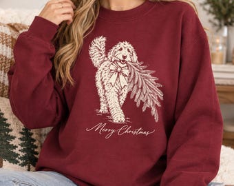 May include: A maroon crewneck sweatshirt featuring a cream-colored line art illustration of a fluffy dog wearing a bow and carrying a pine branch in its mouth. Below the dog, the text Merry Christmas is printed in a cursive script font.