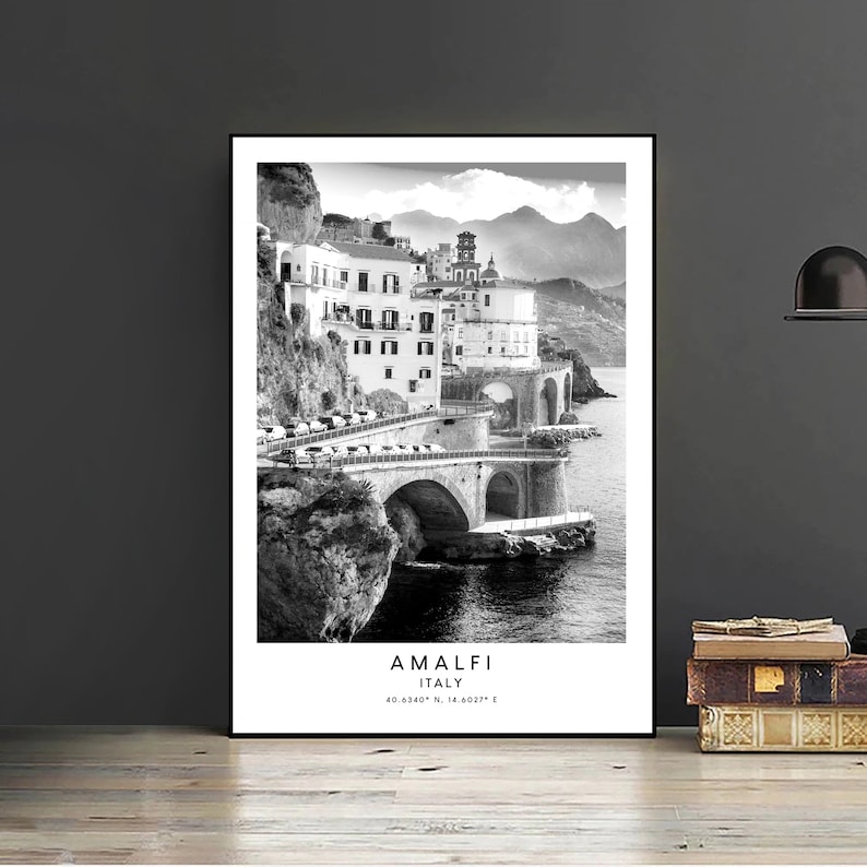 May include: Black and white photograph of a coastal town in Italy. The town is built on a cliffside with a bridge connecting the town to the mainland. The photo includes the text "Amalfi Italy 40.0524&deg; N, 14.6027&deg; E".