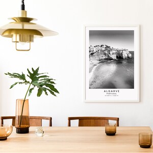 May include: Black and white photograph of a beach in Algarve, Portugal. The photo shows a rocky cliff with buildings on top, a sandy beach, and the ocean. The text "ALGARVE PORTUGAL 37.0188&deg; N, 7.9308&deg; W" is printed below the image.