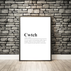 May include: A black and white print with the word "Cwtch" defined as a Welsh word meaning cuddle, hug, or snuggle. The print is framed in a black frame and is hanging on a brick wall.