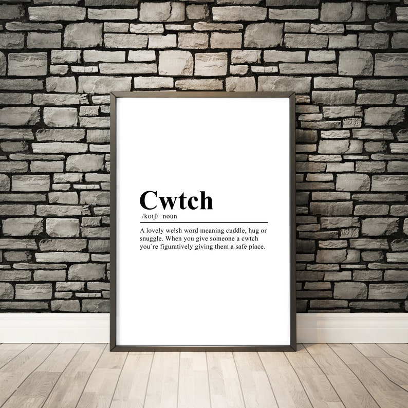 May include: A black and white print with the word "Cwtch" defined as a Welsh word meaning cuddle, hug, or snuggle. The print is framed in a black frame and is hanging on a brick wall.