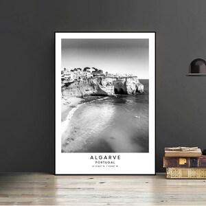 May include: Black and white photograph of a rocky coastline with a beach and buildings in the distance. The text "ALGARVE PORTUGAL 37.0180&deg; N, 7.9208&deg; W" is printed below the image.