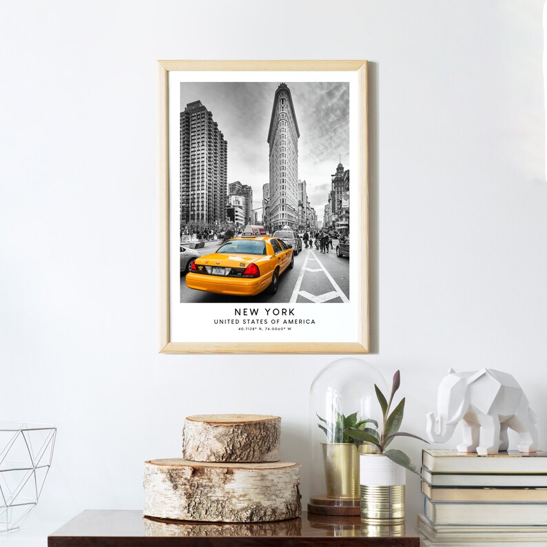 May include: Black and white photograph of a yellow taxi cab driving down a street in New York City. The Flatiron Building is in the background. The text "NEW YORK, UNITED STATES OF AMERICA, 40.7128&deg; N, 74.0060&deg; W" is printed below the image.
