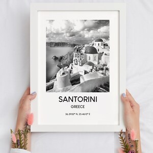 Puede incluir: Fotograf&iacute;a en blanco y negro de Santorini, Grecia. La imagen muestra una ciudad con edificios blancos y c&uacute;pulas azules. El texto "SANTORINI" y "GREECE" est&aacute; impreso debajo de la imagen, junto con las coordenadas "36.3932&deg; N, 25.4615&deg; E".