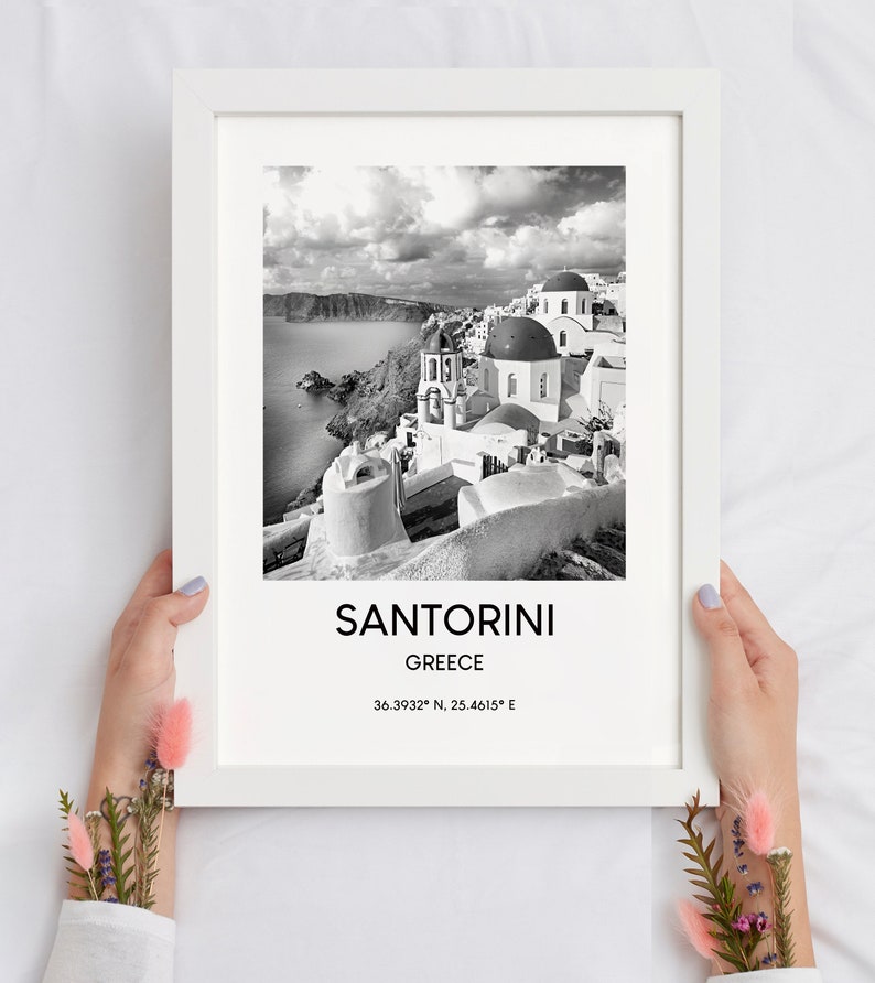 Puede incluir: Fotograf&iacute;a en blanco y negro de Santorini, Grecia. La imagen muestra una ciudad con edificios blancos y c&uacute;pulas azules. El texto "SANTORINI" y "GREECE" est&aacute; impreso debajo de la imagen, junto con las coordenadas "36.3932&deg; N, 25.4615&deg; E".