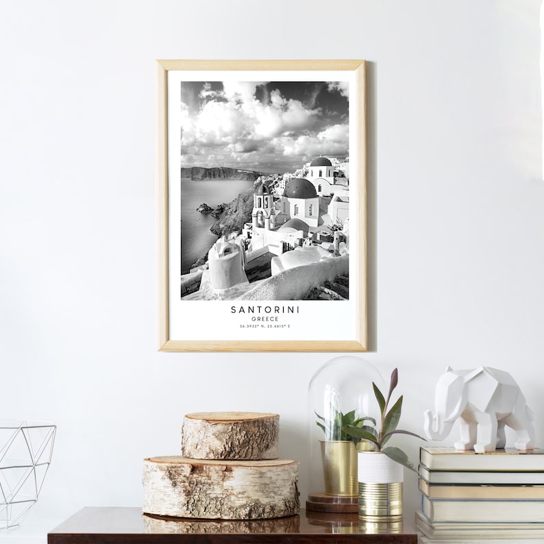 Puede incluir: Fotograf&iacute;a en blanco y negro de Santorini, Grecia. La imagen muestra edificios blancos con c&uacute;pulas azules, una costa rocosa y un cielo azul con nubes blancas. El texto "Santorini Grecia" est&aacute; impreso debajo de la imagen.