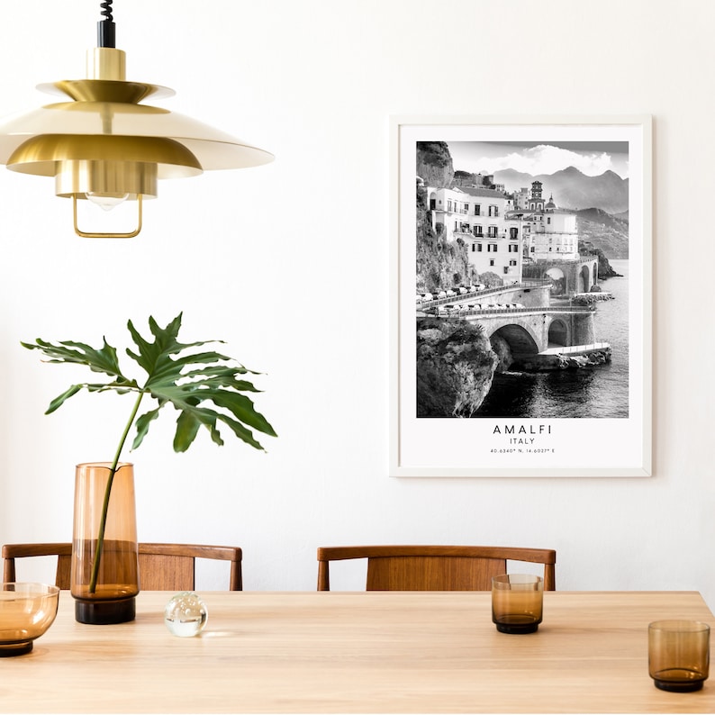 May include: Black and white photograph of the Amalfi Coast in Italy. The photo shows a coastal town with buildings on a cliff overlooking the ocean. The text "Amalfi, Italy 40.6330&deg; N, 14.4037&deg; E" is printed below the image.