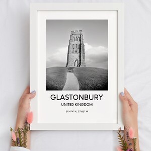 May include: Black and white photograph of Glastonbury Tor, a historic tower in Glastonbury, United Kingdom. The image shows the tower on a hill with a path leading up to it. The text below the image reads "Glastonbury, United Kingdom 51.1474&deg; N, 2.7185&deg; W".