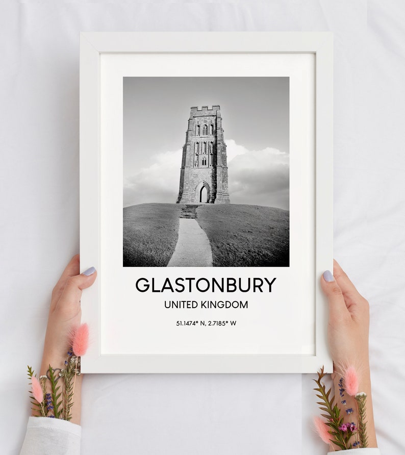 May include: Black and white photograph of Glastonbury Tor, a historic tower in Glastonbury, United Kingdom. The image shows the tower on a hill with a path leading up to it. The text below the image reads "Glastonbury, United Kingdom 51.1474&deg; N, 2.7185&deg; W".