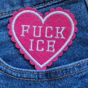 May include: A pink heart-shaped patch with white text that reads "FUCK ICE". The patch has a scalloped edge and is sewn onto a blue denim pocket.