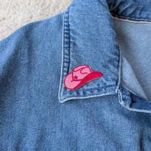 May include: A close-up of a light blue denim jacket collar. A pink cowboy hat patch with a star design is sewn onto the jacket. The hat patch is outlined in a darker pink colour.