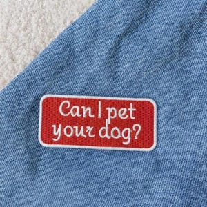 May include: A rectangular embroidered patch with a red background and a white border attached to light blue denim fabric. The patch features the white embroidered text Can I pet your dog? in a cursive font, centered on the red surface.