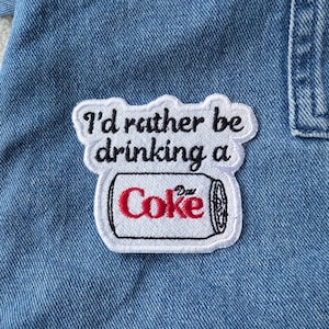 Pode incluir: Patch bordado branco com a frase "I'd rather be drinking a Coke" e uma ilustração de uma lata de Coca-Cola. O patch está costurado em tecido de ganga azul.