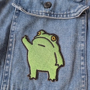 Könnte beinhalten: Nahaufnahme eines hellblauen Jeanshemdes mit einer Tasche. Ein grüner Froschaufnäher mit gelben Augen und schwarzer Umrandung ist auf die Tasche genäht. Der Frosch winkt mit dem Arm.