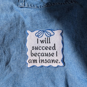 May include: Light blue denim shirt with a white patch embroidered with the phrase "I will succeed because I am Insane." The patch has a blue scalloped border and a bow design at the top.