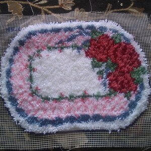 May include: A white oval rug with a pink and blue border and a floral design featuring red roses. The rug is on a white mesh canvas.