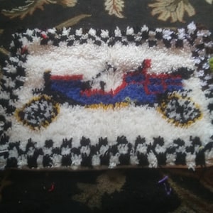May include: Handmade rug featuring a central design of a blue, red, and yellow vehicle on a white background. The rug has a black and white checkered border and yellow wheels. The rug is rectangular in shape.