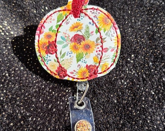 May include: A floral pumpkin-shaped badge reel with a white background and a colorful pattern of sunflowers and red roses. The reel has a red stem and a clear plastic retractable clip with a decorative button.