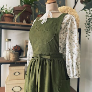 Puede incluir: Un delantal de lino verde con falda fruncida, dos bolsillos grandes y un cinturón de atar. El delantal tiene un detalle curvo en los bolsillos delantero y trasero.