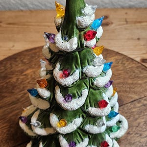 May include: A glossy green ceramic Christmas tree figurine featuring textured white snow accents on the branches. The tree is decorated with numerous small, multi-colored translucent plastic light bulbs in red, blue, yellow, and clear.