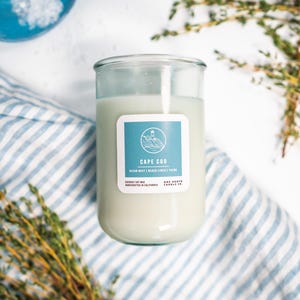 May include: A glass jar candle with a light green wax. The label reads "Cape Cod" with a lighthouse illustration. The candle is on a blue and white striped cloth, with sprigs of thyme and a blue bowl in the background.