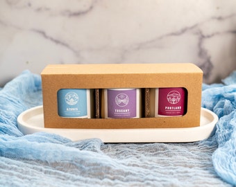May include: Three small glass jars with brown lids and labels. The labels are white with blue text and a small graphic of a city skyline. The jars are arranged in a cardboard box with a white background. The labels read "AZORES", "TUSCANY", and "PORTLAND".