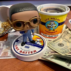 Peut inclure: Une figurine Funko Pop d'un homme portant des lunettes et un costume se tient sur un récipient de Fry Batter. Une tasse d'argent et un récipient de Fry Batter Los Pollos Hermanos sont visibles. Une montre et une chaîne en or sont également présentes, ainsi que diverses coupures de devises américaines.