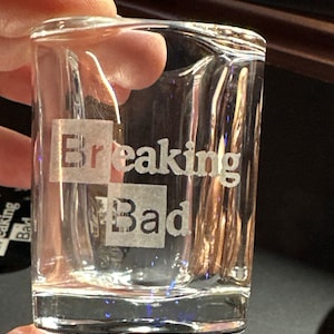 Puede incluir: Vaso de chupito de vidrio transparente con las palabras "Breaking Bad" grabadas. El texto está en negrita, sin serifa. El vaso de chupito es rectangular con una base gruesa.