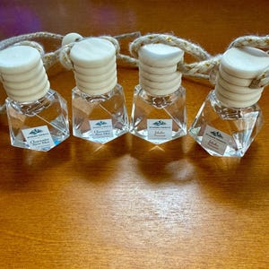 May include: Four clear glass car air fresheners with wooden caps and rope hangers. Each bottle has a label with text. The bottles are arranged on a wooden surface.