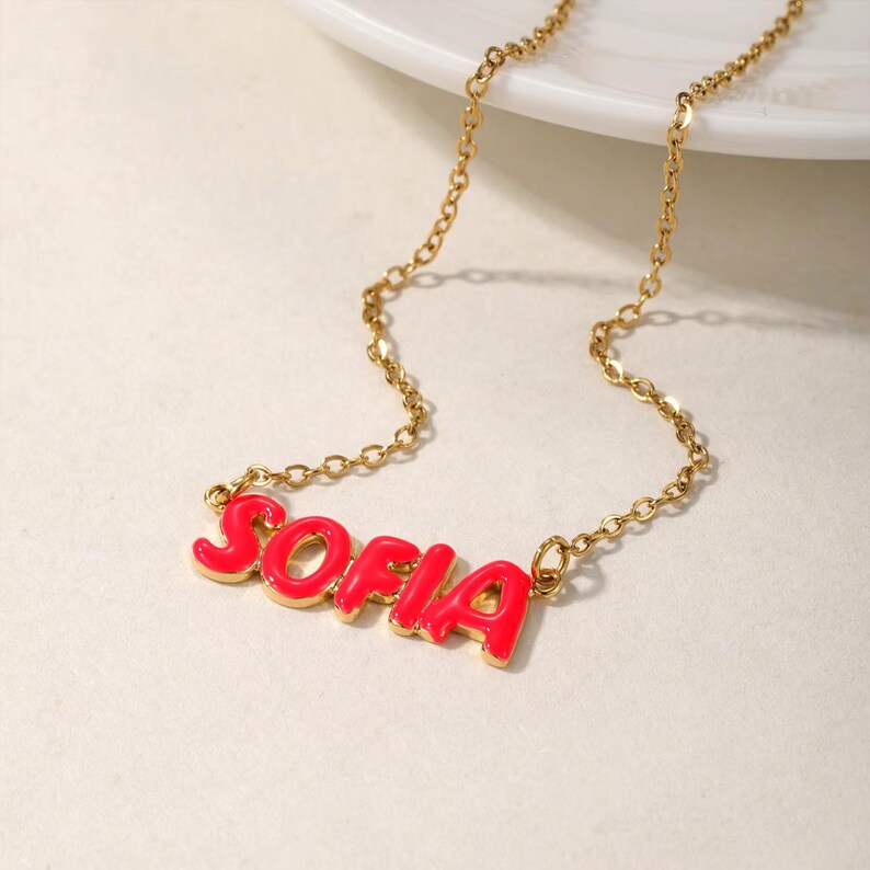 K&ouml;nnte beinhalten: Eine goldene Kettenkette mit einem roten Emaille-Anh&auml;nger, auf dem "SOFIA" steht.