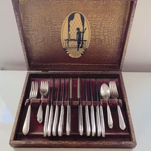 May include: A vintage cutlery set presented in a wooden case. The case has a textured, golden-brown exterior and a maroon velvet interior. The set includes forks, knives, and spoons. The case lid features a silhouette of a person.