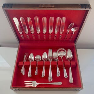 May include: A vintage silver cutlery set presented in a wooden case with a red velvet lining. The set includes knives, forks, spoons, and serving implements. The cutlery is silver-coloured and neatly arranged within the case.