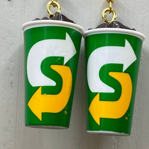 May include: A pair of earrings shaped like a green and white cup with a yellow arrow logo. The logo says "S" with an arrow pointing to the right. The earrings have gold hooks.