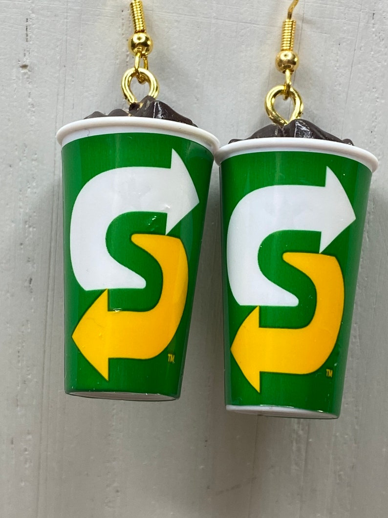 May include: A pair of earrings shaped like a green and white cup with a yellow arrow logo. The logo says "S" with an arrow pointing to the right. The earrings have gold hooks.
