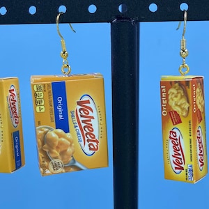 May include: Four pairs of earrings featuring miniature boxes of Velveeta Shells & Cheese. The boxes are yellow and orange with the Velveeta logo and the words "Original" and "Shells & Cheese".