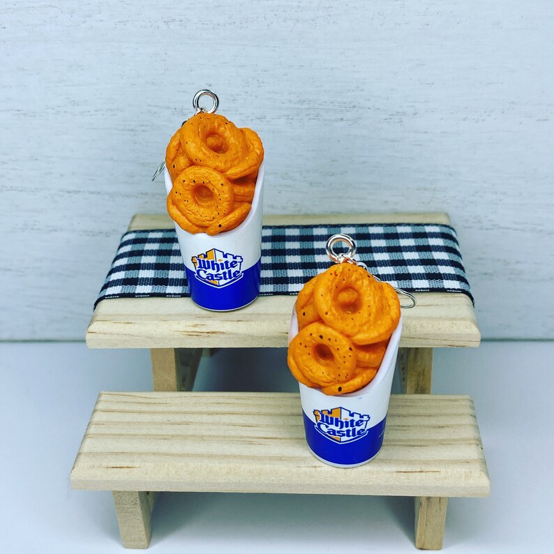 May include: A pair of earrings featuring White Castle onion rings in a small white and blue container. The earrings are on a wooden table with a black and white checkered tablecloth.