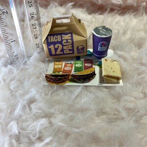 May include: A miniature Taco Bell 12 pack box, a purple cup with the Taco Bell logo, and three miniature tacos with different toppings on a white surface. The tacos have labels that say "Mild", "Hot", "Verde", and "Fire".