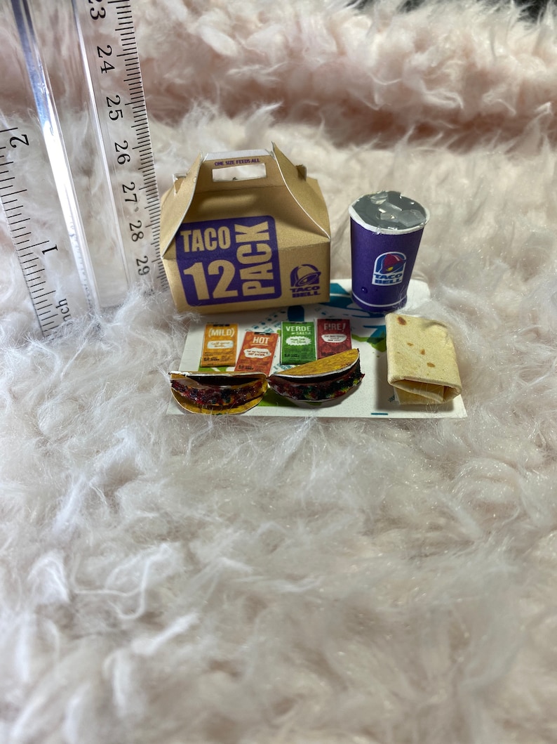 May include: A miniature Taco Bell 12 pack box, a purple cup with the Taco Bell logo, and three miniature tacos with different toppings on a white surface. The tacos have labels that say "Mild", "Hot", "Verde", and "Fire".