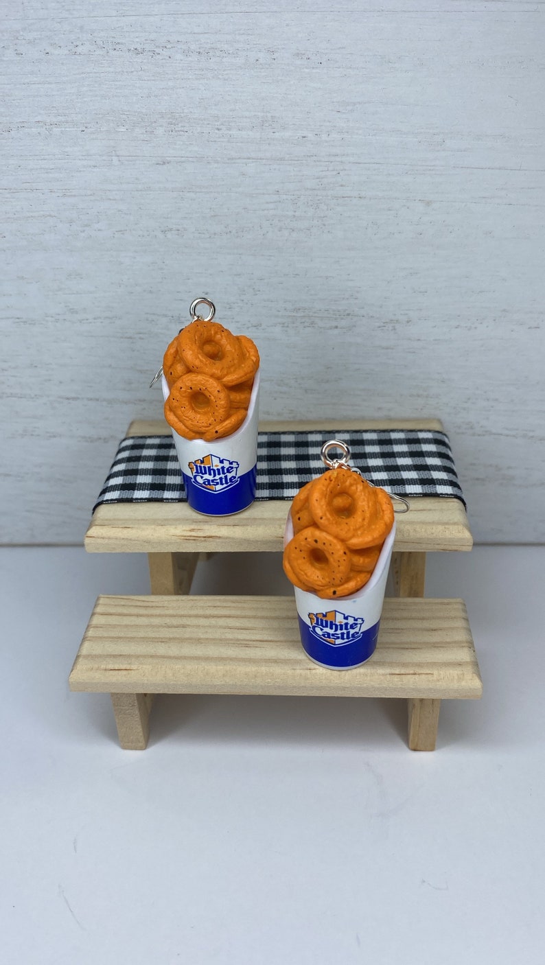 May include: A pair of earrings featuring White Castle onion rings in a blue and white paper cup. The earrings are on a wooden table.