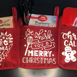 May include: Three red oven mitts with white text and designs. The left mitt has a heart-shaped design with the words "Milk, Cocoa, Sugar, Vanilla, Butter, Flour" and "Cinnamon Mom". The middle mitt has a whisk and the words "We whisk you a Merry Christmas". The right mitt has a gingerbread man and the words "Christmas Calories Don't Count".
